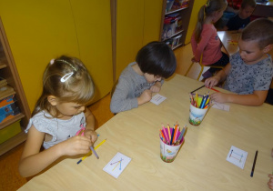 Dwoje dzieci trzyma w ręku kredki i układa je na stoliku według wzoru na kartce. Jeden chłopiec ogląda z zaciekawieniem swoją kartkę z wzorem ułożenia kolorowych kredek.